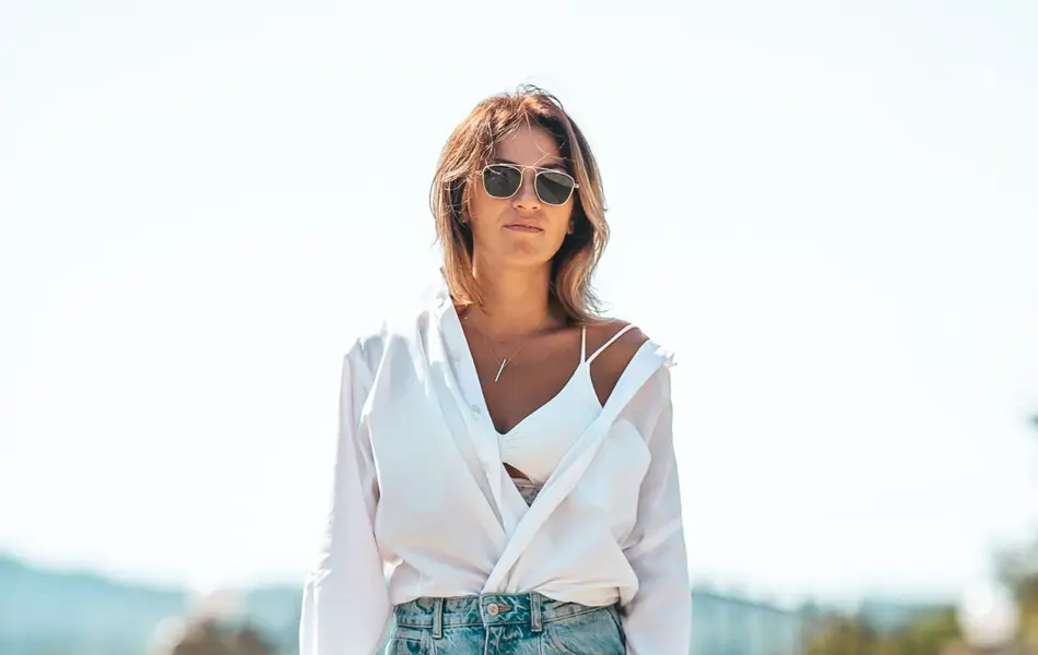 Dame går på grusvei iført hvit åpen bluse, hvit bh, sandaler, solbriller og løse baggy jeans.