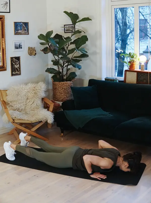 treningsekspert Anna Johansen trener på matte på gulvet, mørkegrønn sofa i bakgrunnen, lys stol med pels oppi, str grønn plante, bilder på veggen
