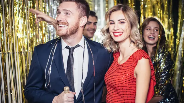 Mann med blå dress og dame med rød paljettkjole står i et festlokale som er pyntet med glitterremser