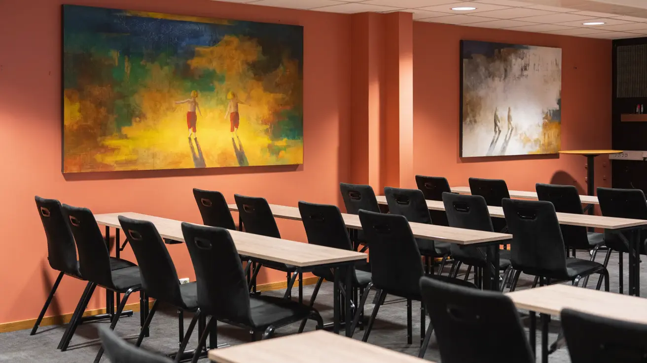 Møterom med klasseroms oppsett, svarte stoler og lyse trebord. Lys rød farge på veggen.