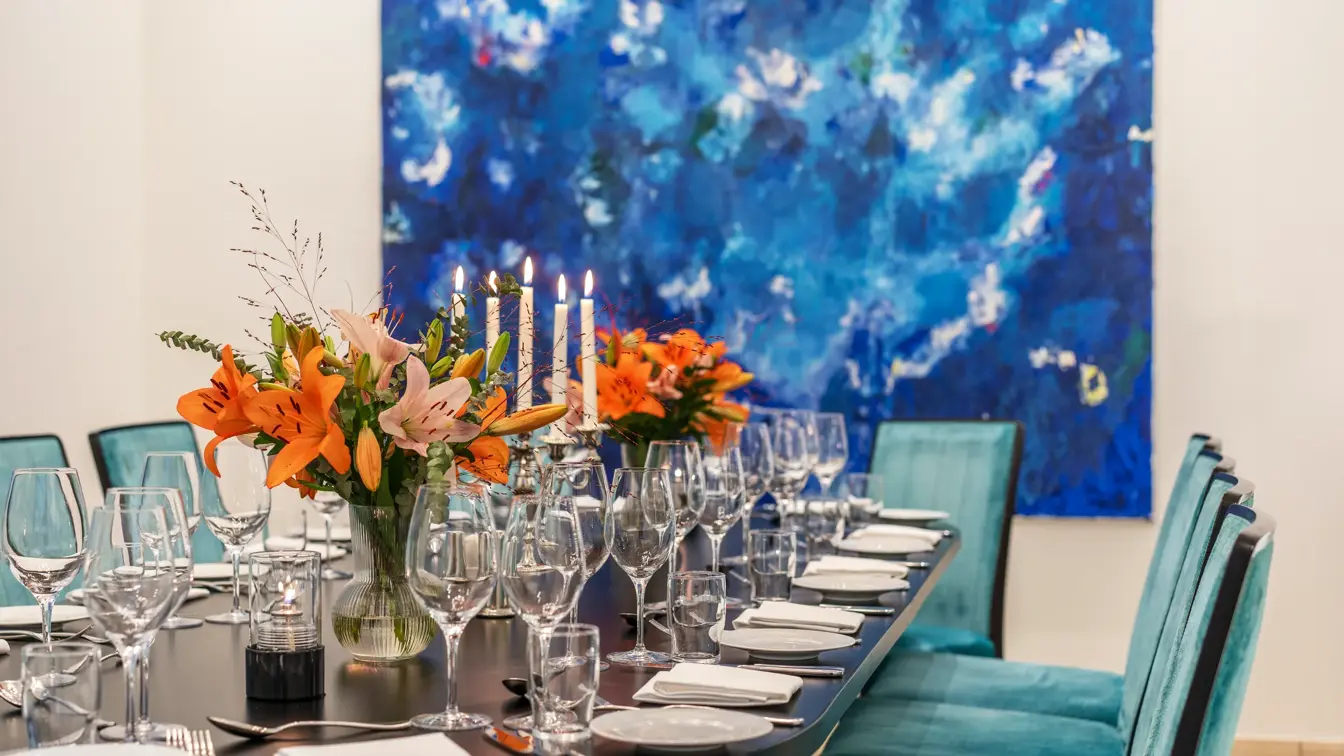 Oppdekket langbord med oransje liljer på bordet, turkise stoler, blått maleri på veggen.