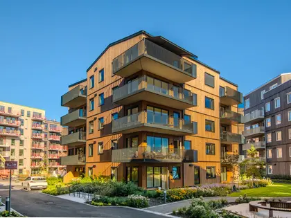 Bildet viser en blokk med leiligheter i boligprosjektet Skårerløkka PREMIUM på Lørenskog. Blokken er i lyst tre med balkonger i mørkt materiale. Det er et grønt område rundt blokken og blå himmel.