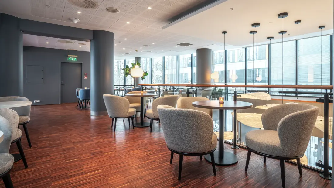 Lounge i 2. etasje med grå hvilestoler rundt runde bord. Parkettgulv. Vinduer fra tak og ned til gulvet i 1. etasje.