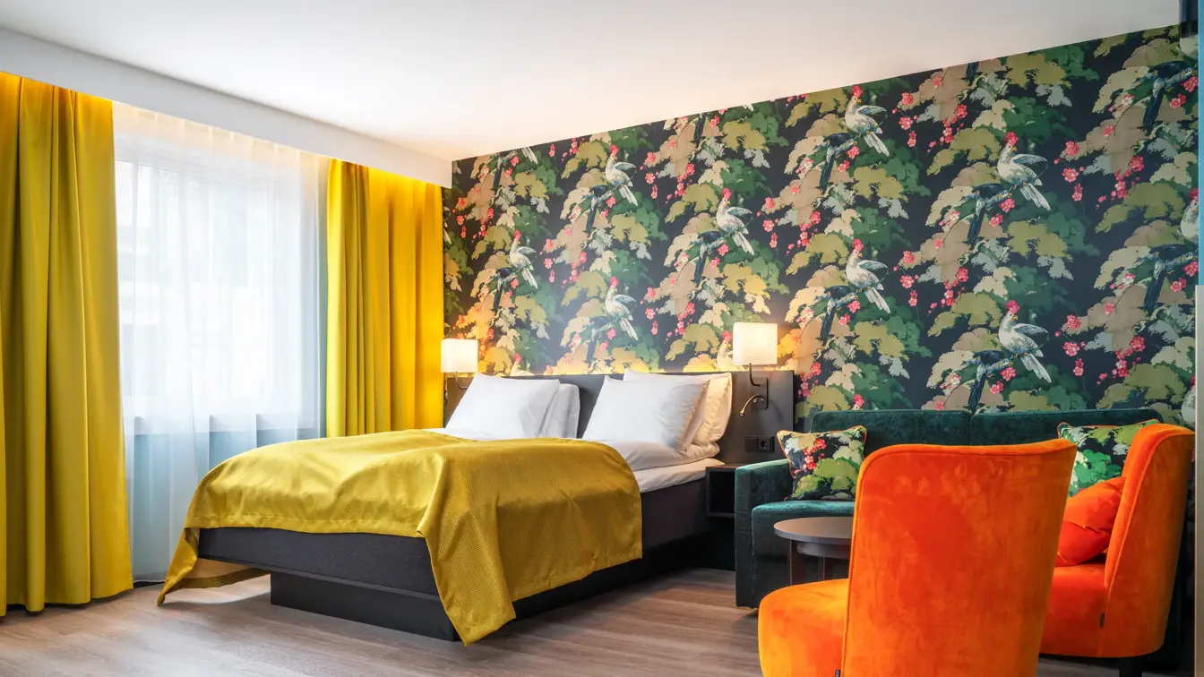 Vindu med gule gardiner. Seng med gult sengeteppe. Fargerik tapet med fugler og blomster, grønn sofa, oransje stoler og mørkt bord i business rom på Thon Hotel Bristol Bergen