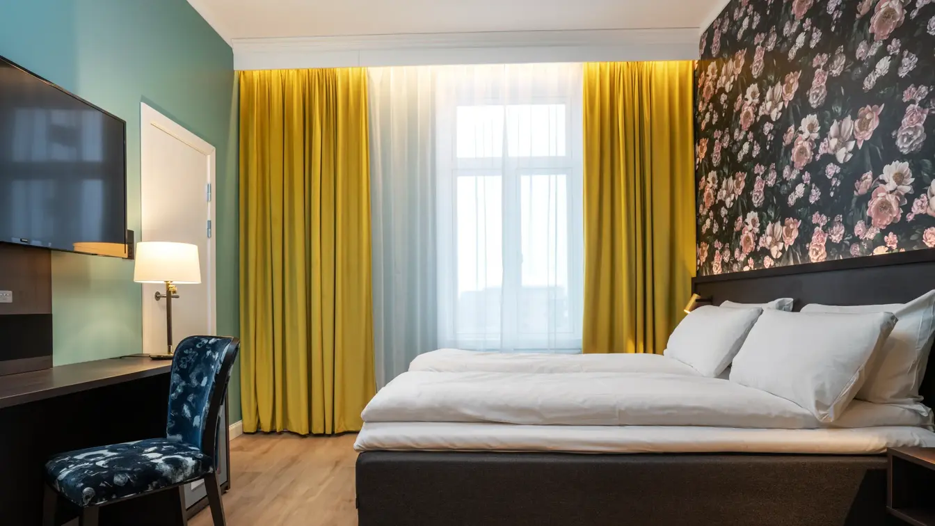 Stort vindu med gule gardiner fra tak til gulv. Mørk tapet med store lyserosa roser. Dobbeltseng. Mørkt skrivebord med veggehengt TV over og mønstrete stol.  I dobbeltrom på Thon Hotel Høyers i Skien.