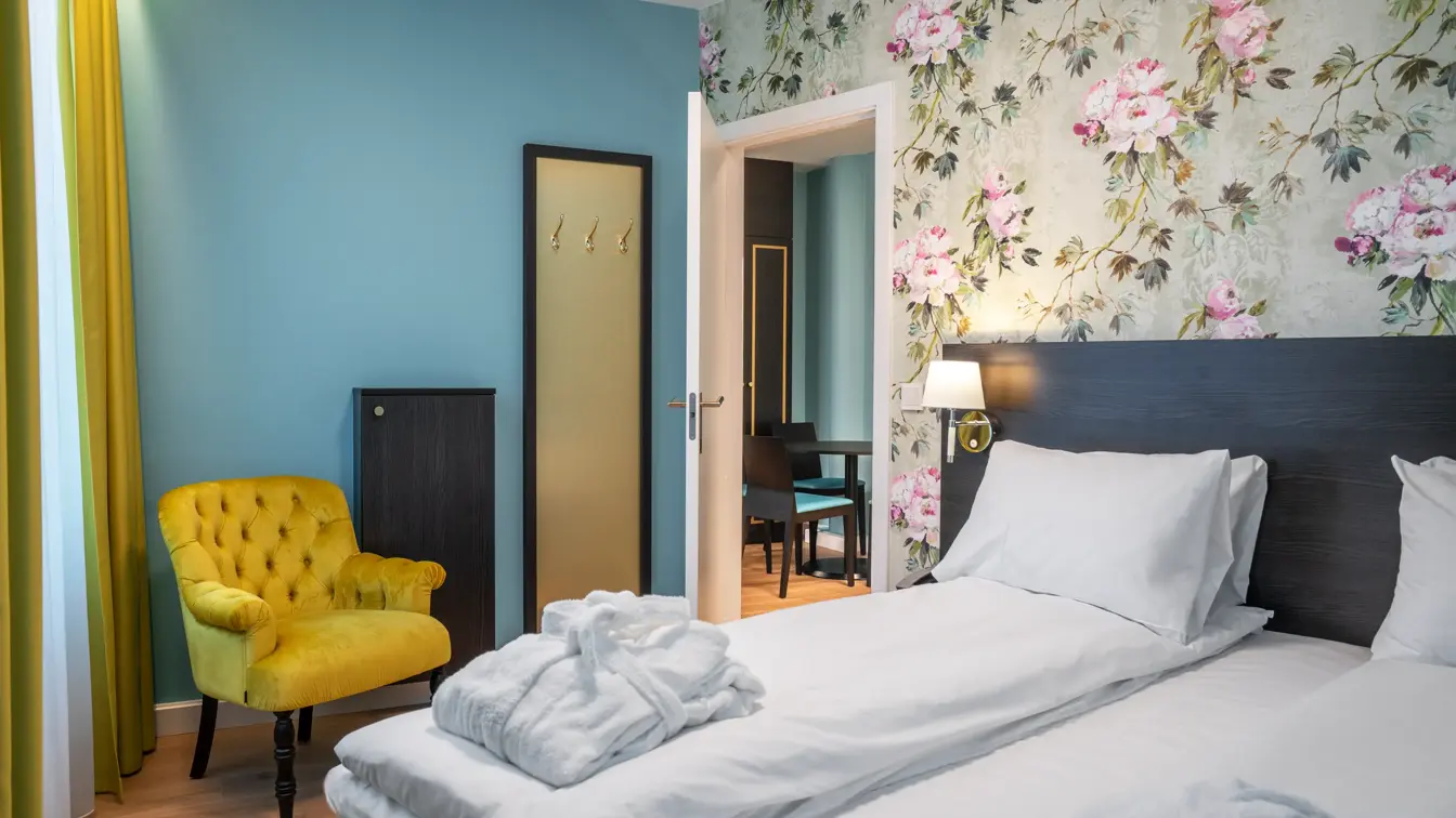 Blomstrete tapetvegg, dobbeltseng, blå vegg. gule gardiner, gul stol, speil på vegg, vindu fra gulv til tak. 