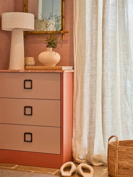 Rosa kommode med lampe og vase, gardiner og speil på veggen.