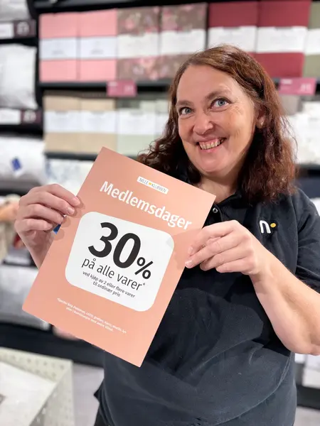 Mørkhåret dame i mørk t-shirt holder en beige og hvit plakat, med fargerike gardinpakker bak seg