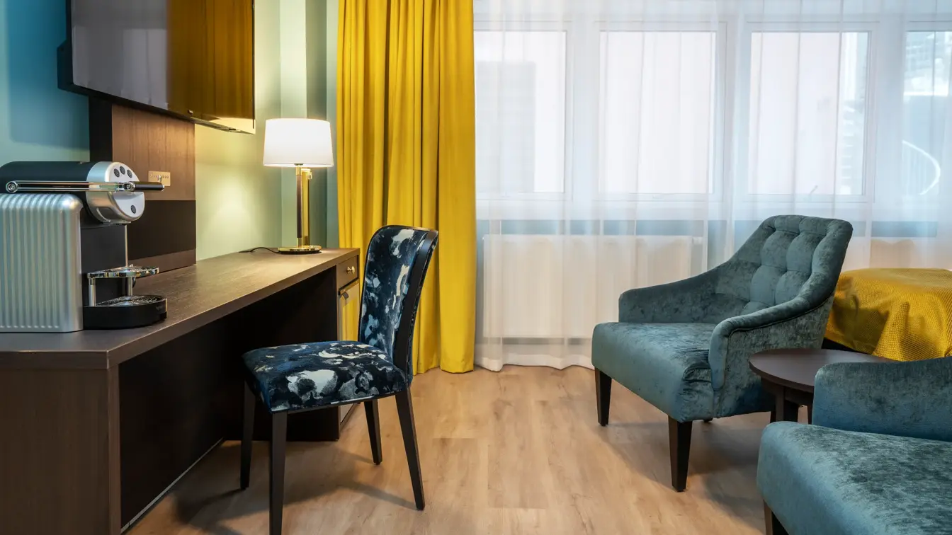 Skrivebord med kaffemaskin, vegghengt TV over og mørk mønstret stol. Vinduer og gule gardiner fra tak til gulv. To turkise lenestoler. I superior rom på Thon Hotel Høyers i Skien.