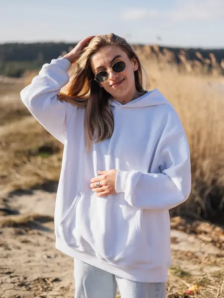 Dame med hvit hettegenser og solbriller poserer foran åker.  