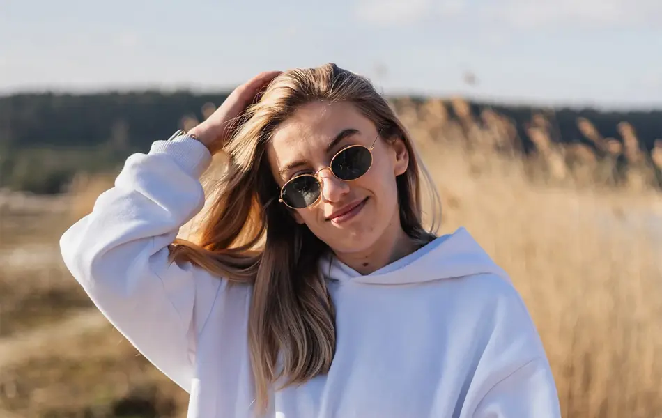 Dame med hvit hettegenser og solbriller poserer foran åker.  