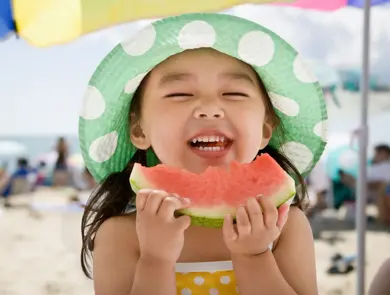 Jente med hatt spiser melon på stranda