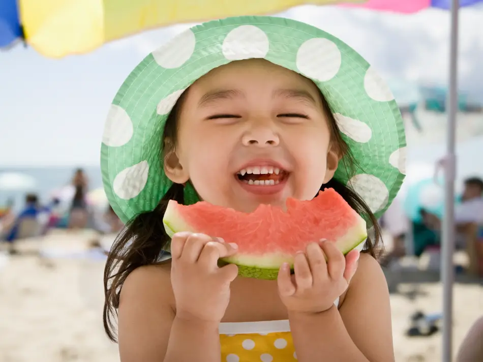 Jente med hatt spiser melon på stranda