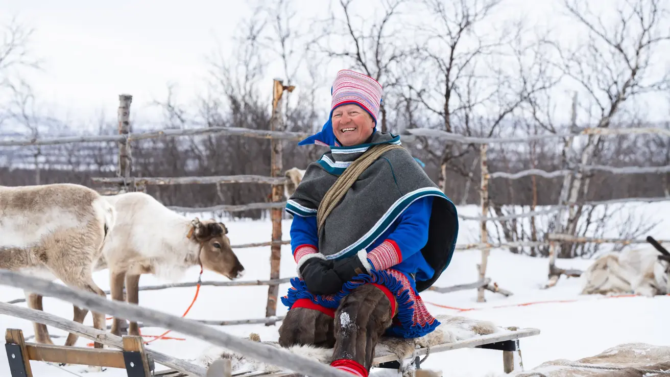Smilende mann i samekofte sitter på en slede ved siden av et reinsdyr