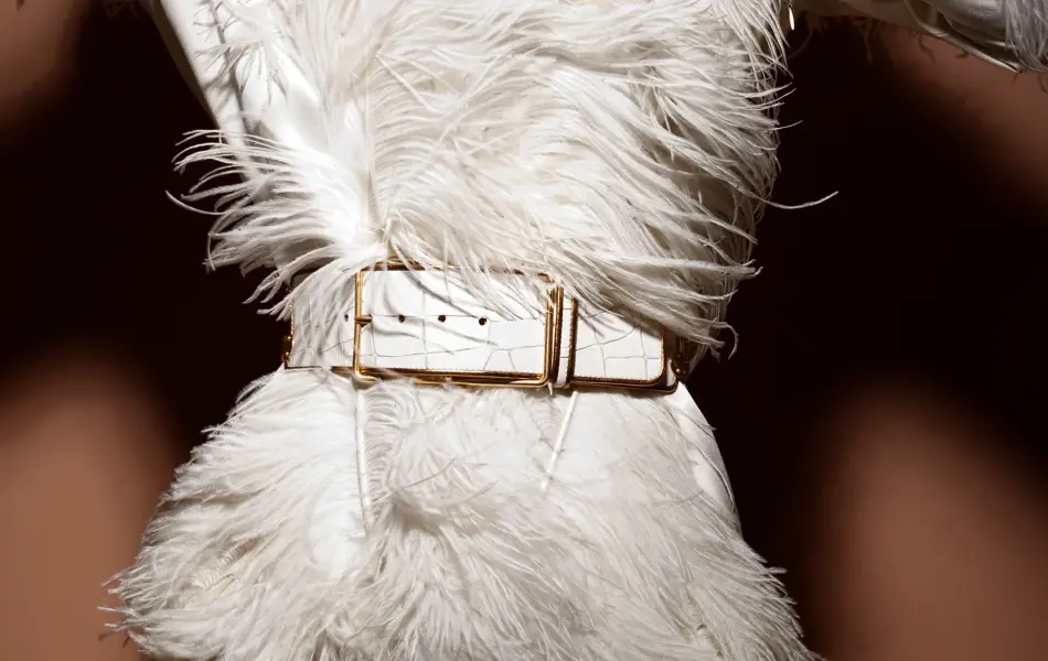 En kvinne poserer mot en brun vegg. Hun er iført en hvit kjole med hvit fjærkreasjon og et hvitt belte.