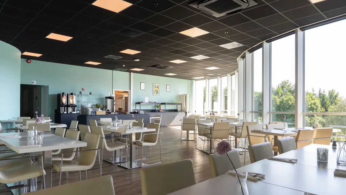 Oversiktsbilde av frokostsalen med bord og stoler, store vinduer med utsikt og buffet bakerst i lokalet.