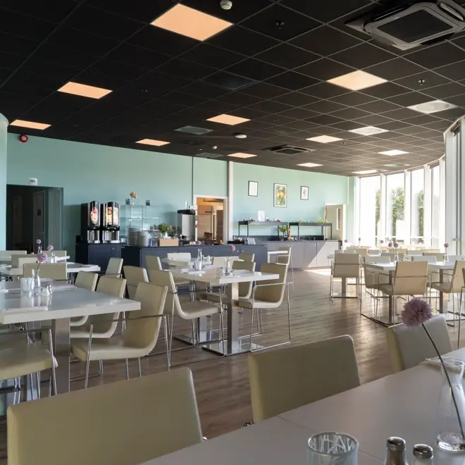 Oversiktsbilde av frokostsalen med bord og stoler, store vinduer med utsikt og buffet bakerst i lokalet.