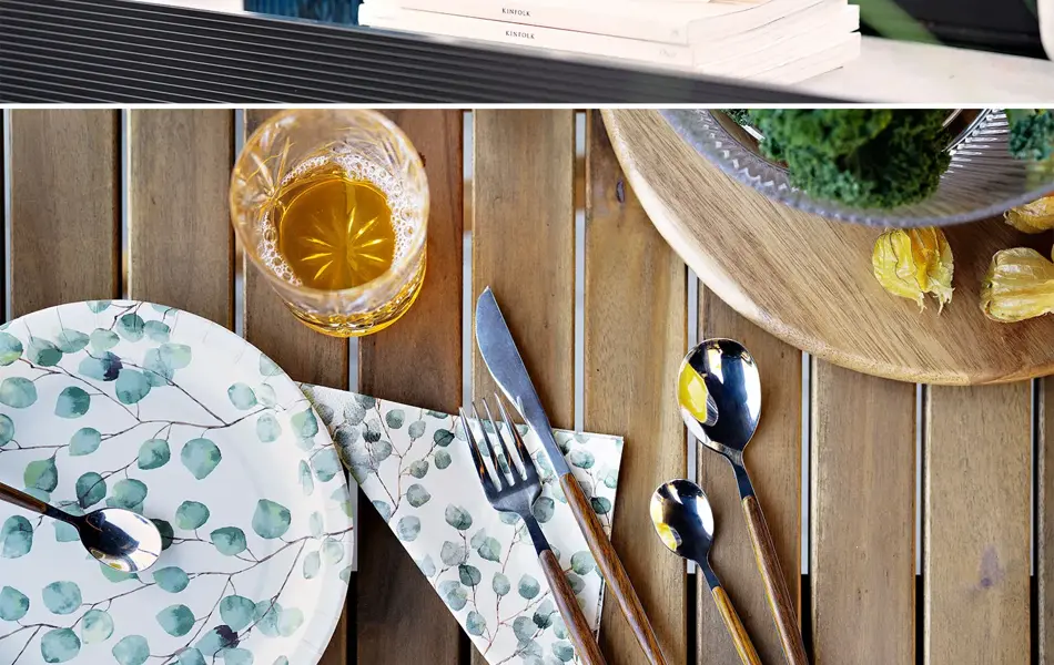 Blå vase med blå blomster, hvit skjellvase og papptallerken med bestikk på et bord.