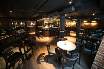 Brune bord og stoler, runde, lyse marmorbord med tilhørende brune stoler. I Bakgrunnen sees bardisken med tilhørende, brune barstoler. På veggene henger det TV-er i ramme. TV-ene viser en peis.