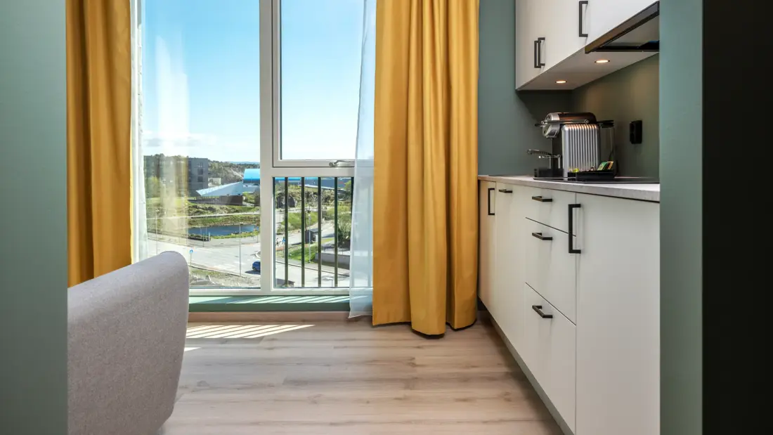 Standard queen rom med kjøkken krok, stort vindu med gule gardiner, parkett og grønne vegger.