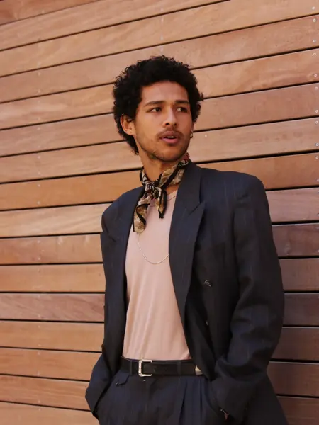 Mann med dress og silketørkle i halsen poserer inntil vegg.