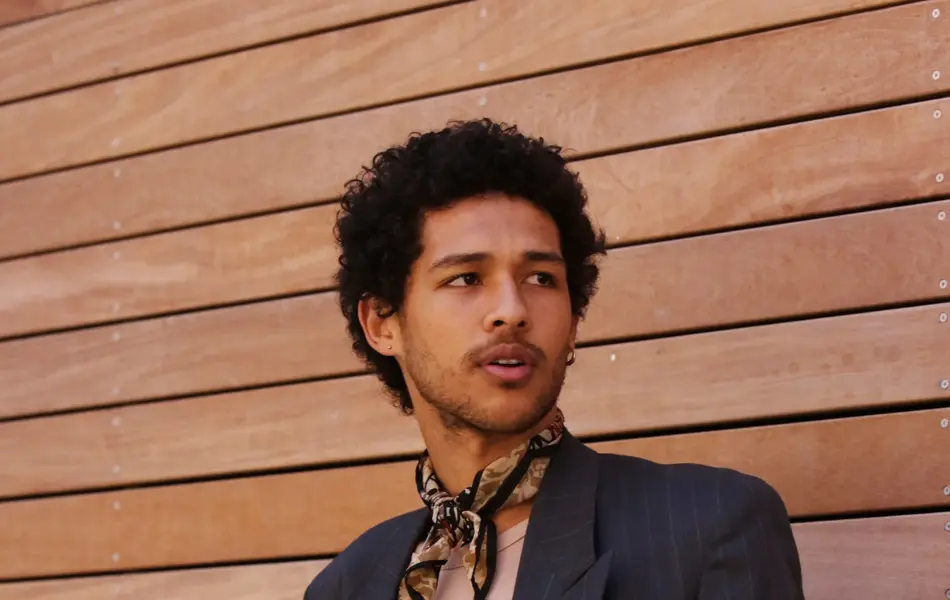 Mann med dress og silketørkle i halsen poserer inntil vegg.