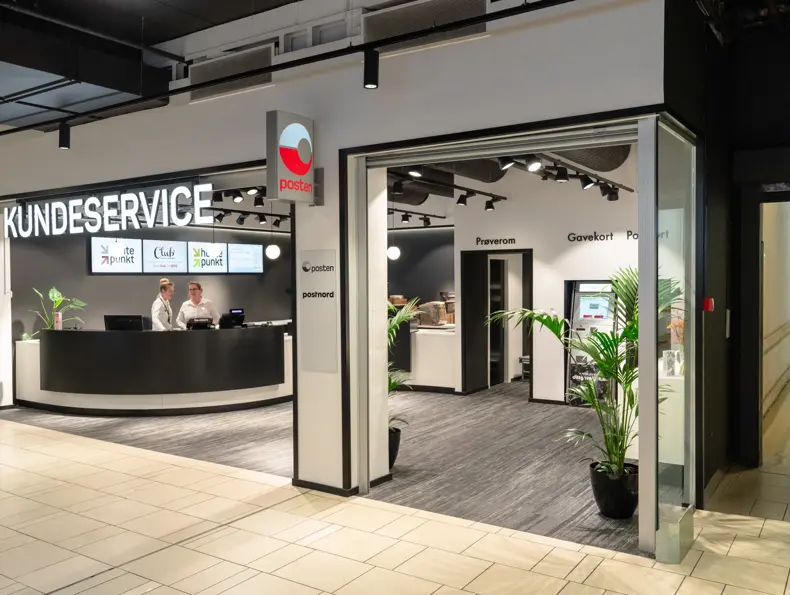 Bilde av Kundeservice lokale med skilt Posten og 2 ansatte bak disken som har 4 digitale skjermer på bakveggen