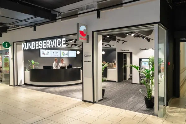 Bilde av Kundeservice lokale med skilt Posten og 2 ansatte bak disken som har 4 digitale skjermer på bakveggen