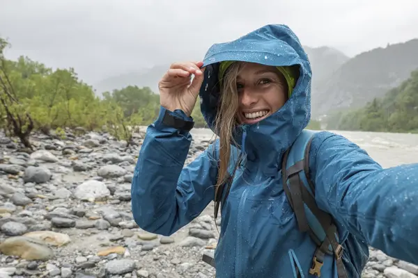 Ung kvinne står ute i regnvær i fjellet med blå allværsjakkke og sekk.