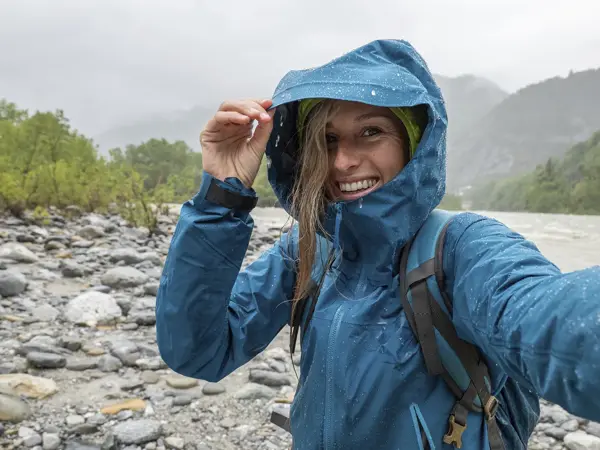 Ung kvinne står ute i regnvær i fjellet med blå allværsjakkke og sekk.