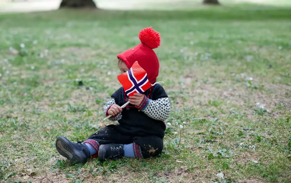 Lite barn i festdrakt sitter på gresset med norsk flagg. Bilde til artikkel om aktiviteter for barn på 17. mai.