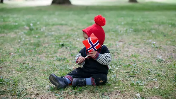 Lite barn i festdrakt sitter på gresset med norsk flagg. Bilde til artikkel om aktiviteter for barn på 17. mai.