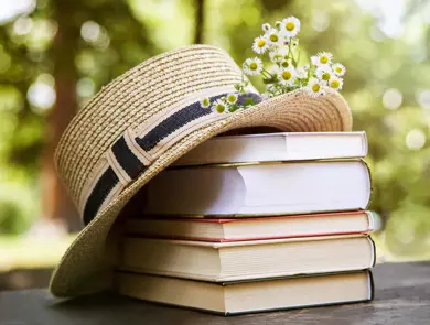 Fem bøker ligger i en stabel på et bord ute, med en stråhatt og noen markblomster på toppen.