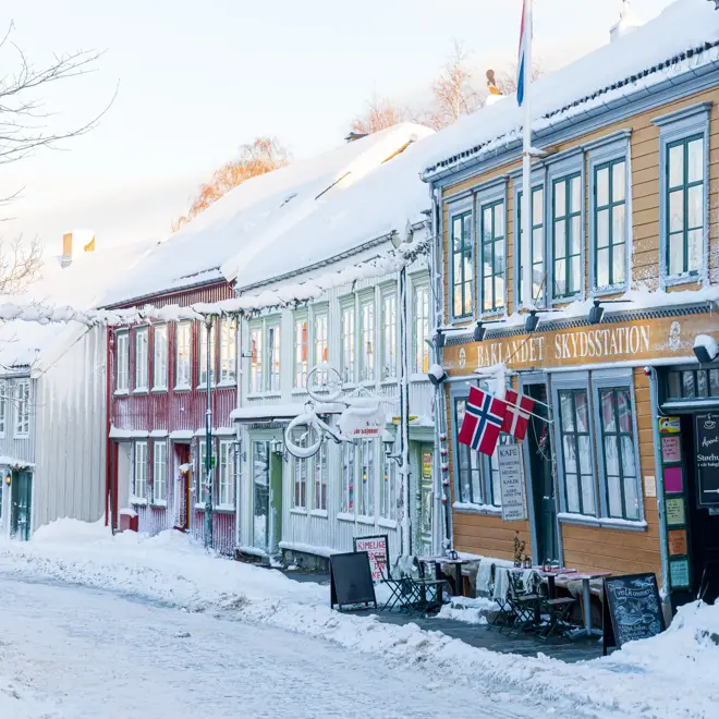Snødekt gate på Bakklandet i Trondheim med fargerike trehus. Norske flagg henger over inngangen til kaféen Bakklandet Skydsstation.