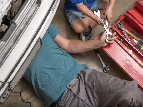 Fugleperspektiv på mann som ligger under bil og reparerer noe, barn gir ham verktøy. Gråtoner. Foto til artikkel om gaver til pappatyper på farsdag. 