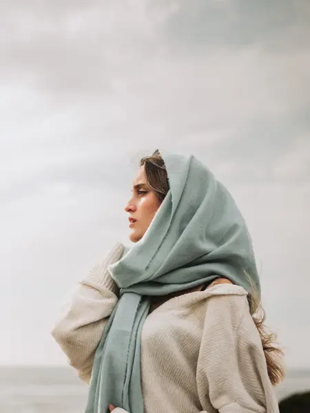 Portrettfoto av dame med stort skjerf rundt hodet og stor genser, ser utover havet, overskyet vær i bakgrunnen.