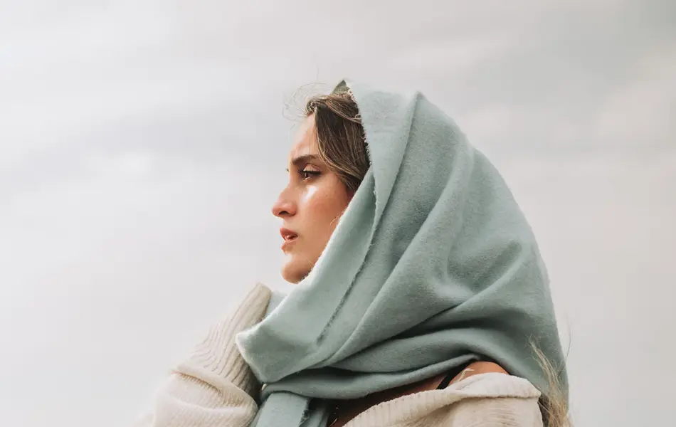 Portrettfoto av dame med stort skjerf rundt hodet og stor genser, ser utover havet, overskyet vær i bakgrunnen.