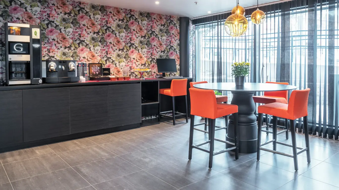 Oransje barstoler, rundt sort bord, fargerik blomstertaper, gjest PC og kaffemaskin i resepsjonen på Thon Hotel Hammerfest
