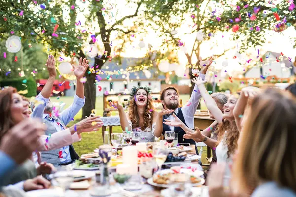 Brudepar sitter ved enden av et utendørs langbord mens gjestene skåler rundt, uskarp kveldssol i bakgrunnen. Foto til artikkel om bryllupsmenyer tilpasset sesong.