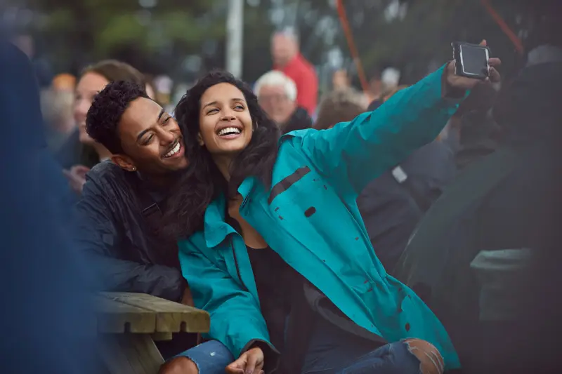 Dame med blå allværsjakke tar selfie med mann på en benk på festival 