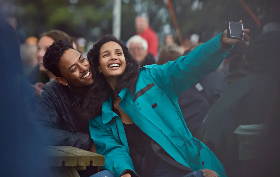 Dame med blå allværsjakke tar selfie med mann på en benk på festival 