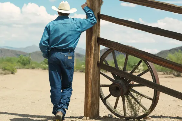 Mann i dobbel denim jeans og denimskjorte og cowboyhatt bakgra på en ranch som titter ut over landskapet.