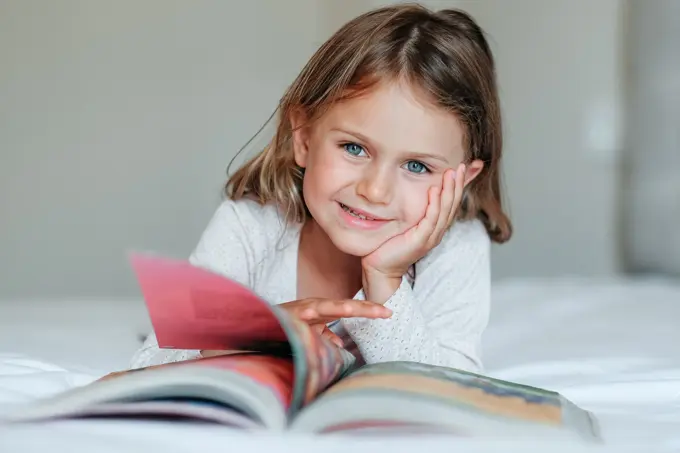 Happy girl kid lies on bed, read the book. Comfort, cozy home concept. Education develop child toddler