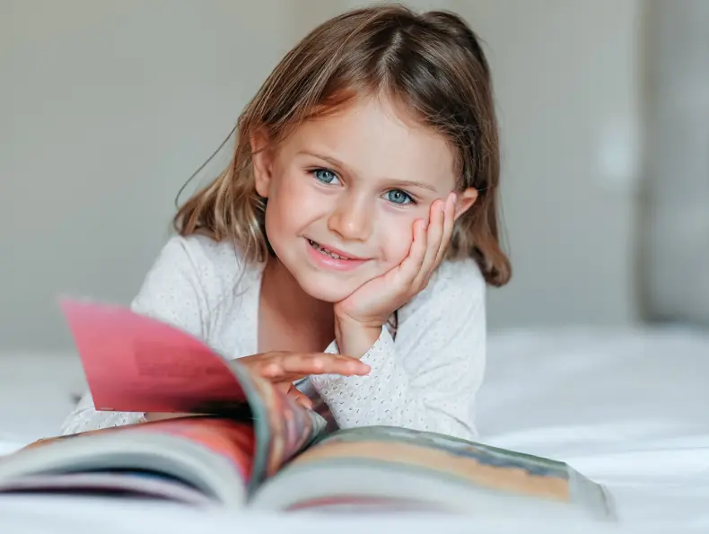 Happy girl kid lies on bed, read the book. Comfort, cozy home concept. Education develop child toddler