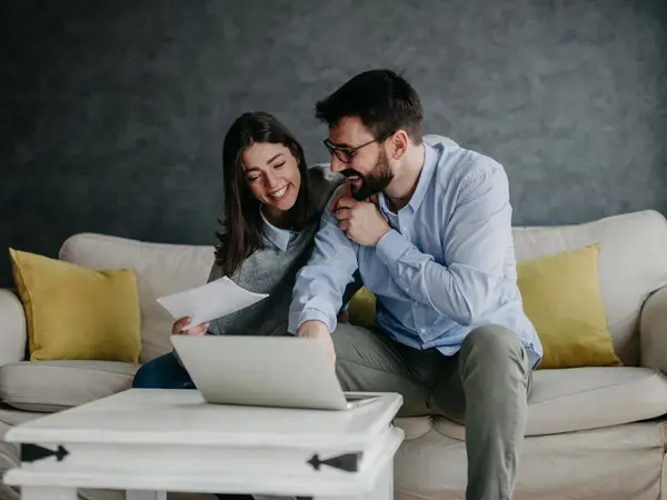 Dame og mann sitter i sofa foran PC og diskuterer, damen holder et ark, mannen peker på noe. Foto til artikkel om sjekkliste til bryllupsplanlegging.