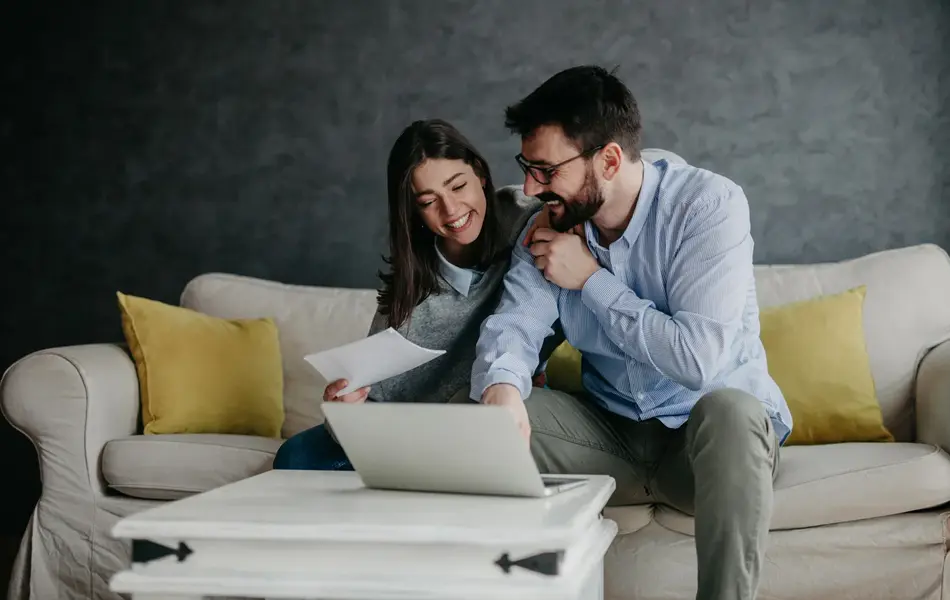Dame og mann sitter i sofa foran PC og diskuterer, damen holder et ark, mannen peker på noe. Foto til artikkel om sjekkliste til bryllupsplanlegging.