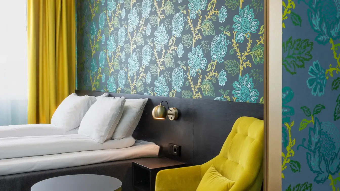 Blå og grønn mønstrete tapet med blomster, gul stol og rundt bord, dobbelsteng og gule gardiner i dobbeltrom på Thon Hotel Kirkenes.