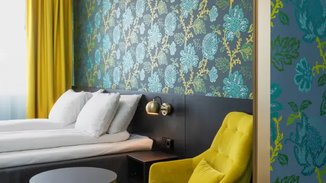 Blå og grønn mønstrete tapet med blomster, gul stol og rundt bord, dobbelsteng og gule gardiner i dobbeltrom på Thon Hotel Kirkenes.