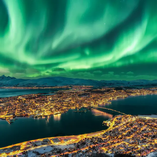 Aurora Borealis dancing over Tromso Urban Skyline.  Tromso Panorama and winter night landscape with Northern Lights (Aurora Polaris) on a cold night, with starry sky, polar lights and snowcapped mountains on Kvaloya Island in the background. Cityscape with Brightly Street Light of the Arctic Capital, Northern Norway