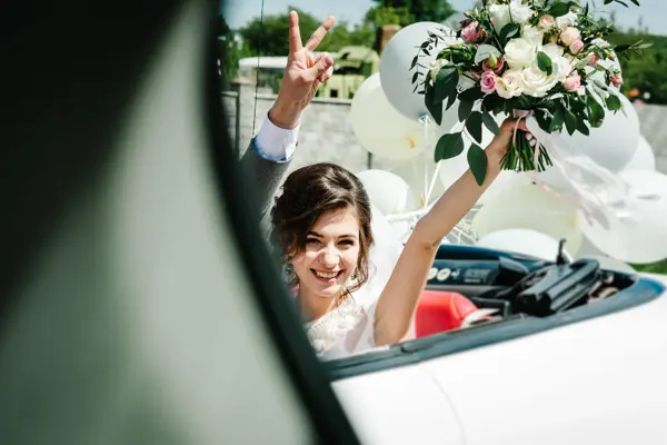 A newlywed wedding couple is driving a convertible retro car with balloons on a country straight road for their honeymoon. Way near spring field of flowers. The best day and marriage. Just married.